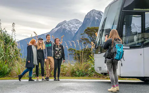 Group posing for a photo with a tour bus and snow-capped mountains in Milford Sound, New Zealand.