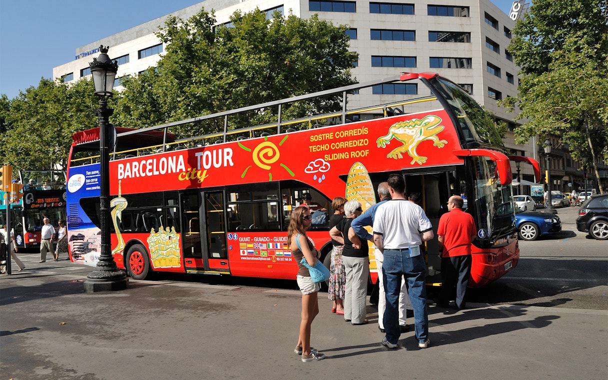 Barcelona hop-on hop-off bus with tourists boarding near city landmarks.