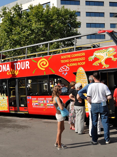 Barcelona hop-on hop-off bus with tourists boarding near city landmarks.