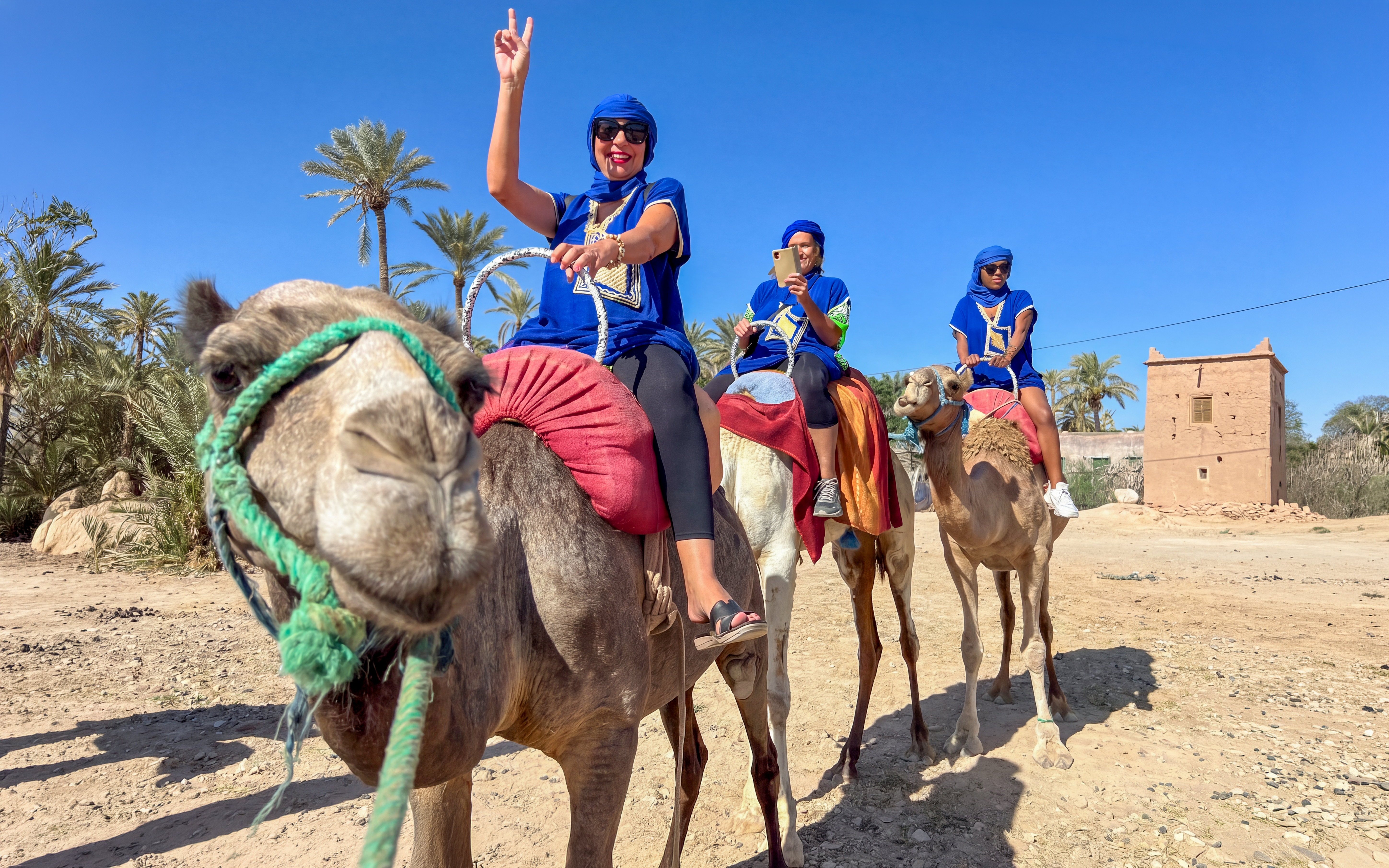 Camel ride in Palmeraie with tourists wearing traditional attire, palm trees and a clay building in the background.