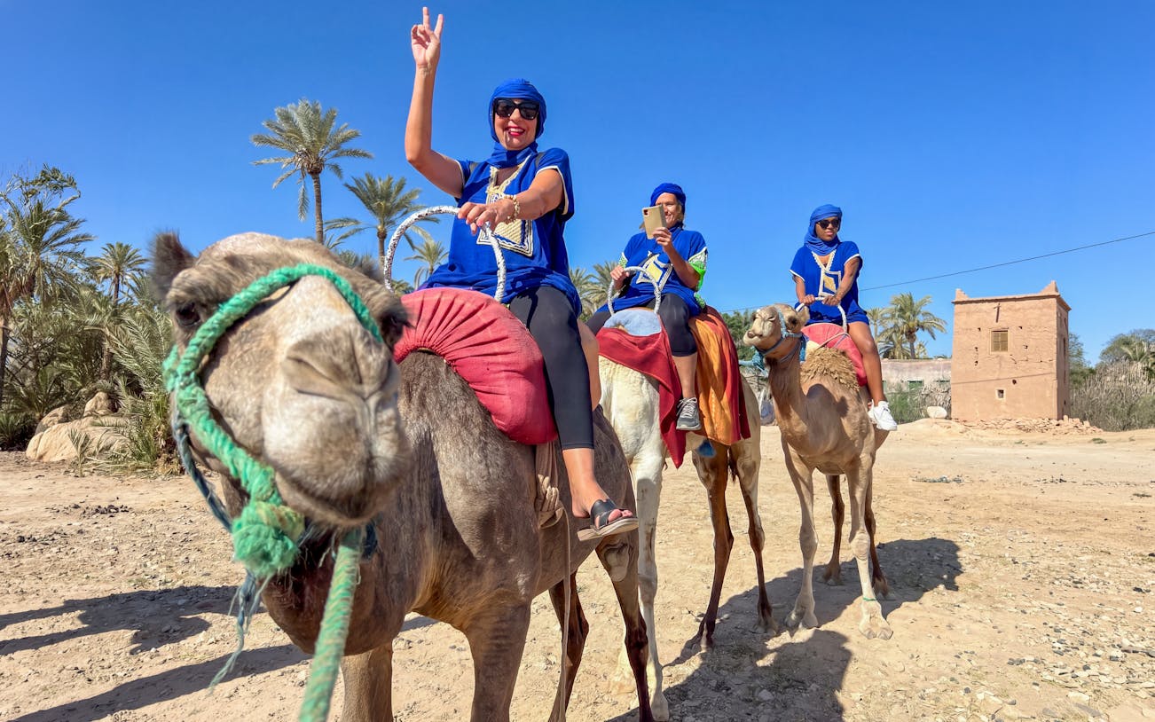 Camel ride in Palmeraie with tourists wearing traditional attire, palm trees and a clay building in the background.