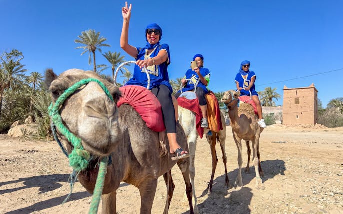 Camel ride in Palmeraie with tourists wearing traditional attire, palm trees and a clay building in the background.