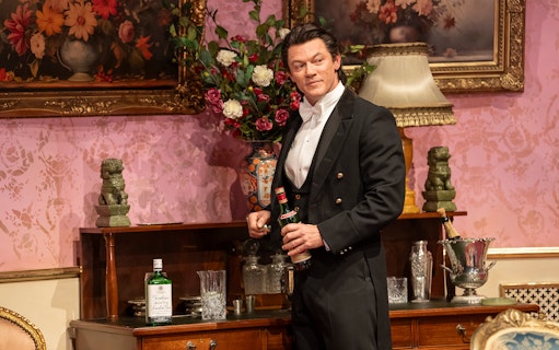 Man in formal attire holding a bottle in an elegant room with floral decor, related to Backstairs Billy.
