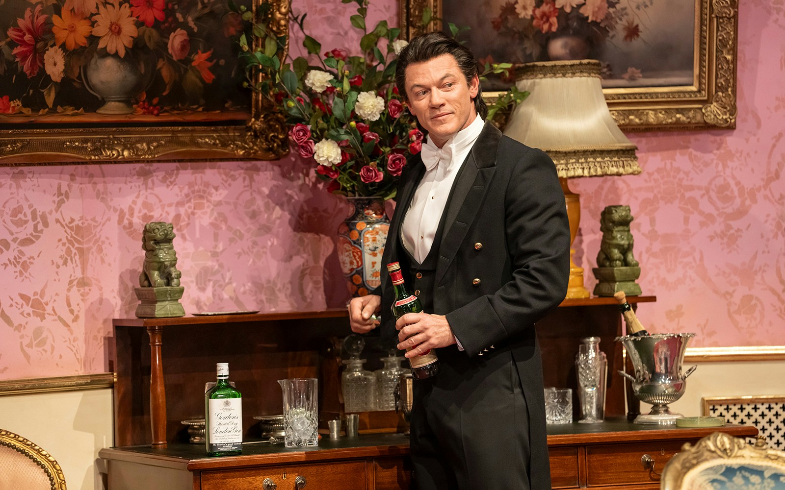 Man in formal attire holding a bottle in an elegant room with floral decor, related to Backstairs Billy.