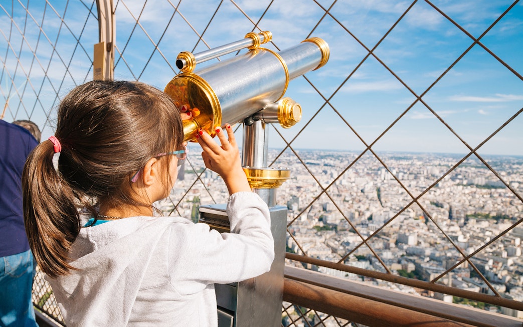 Child using telescope on Eiffel Tower viewing platform, Paris cityscape in background.
