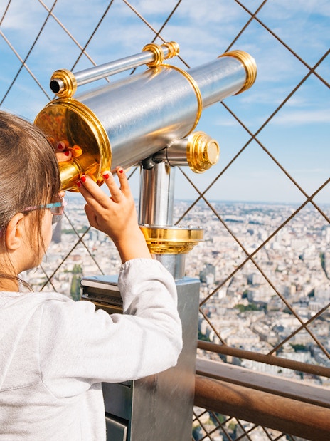 Child using telescope on Eiffel Tower viewing platform, Paris cityscape in background.