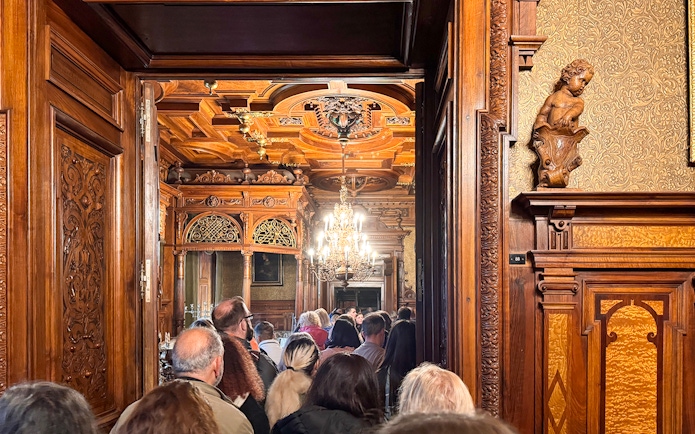 Guests touring ornate interior of Peles Castle with carved wood and chandelier.