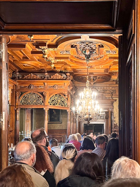 Guests touring ornate interior of Peles Castle with carved wood and chandelier.
