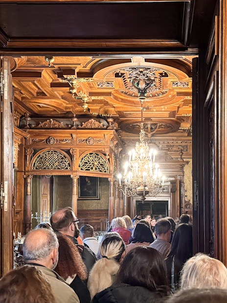 Guests touring ornate interior of Peles Castle with carved wood and chandelier.