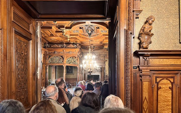 Guests touring ornate interior of Peles Castle with carved wood and chandelier.