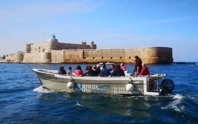 Speed boat near Syracuse sea caves with tourists, castle in background.