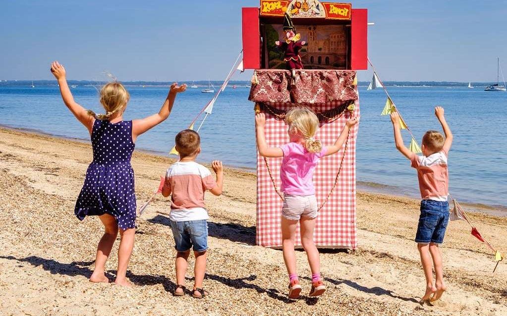 Children watching a puppet show on a sandy beach with sailboats in the background.
