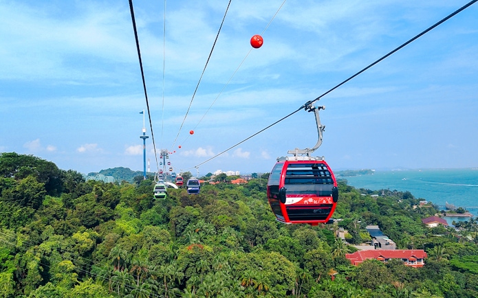 Tourists riding Singapore Cable Car over lush greenery and coastline.