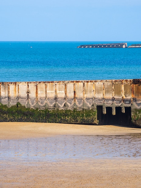 Remains of a D-Day landing craft on the beach in Caen, France, with visitors exploring nearby.