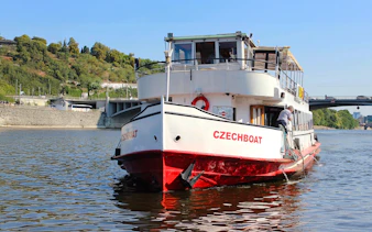 Historic ship Maria Croon on Vltava River, Prague, near a bridge and green hillside.