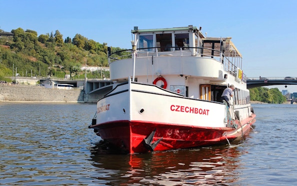 Historic ship Maria Croon on Vltava River, Prague, near a bridge and green hillside.