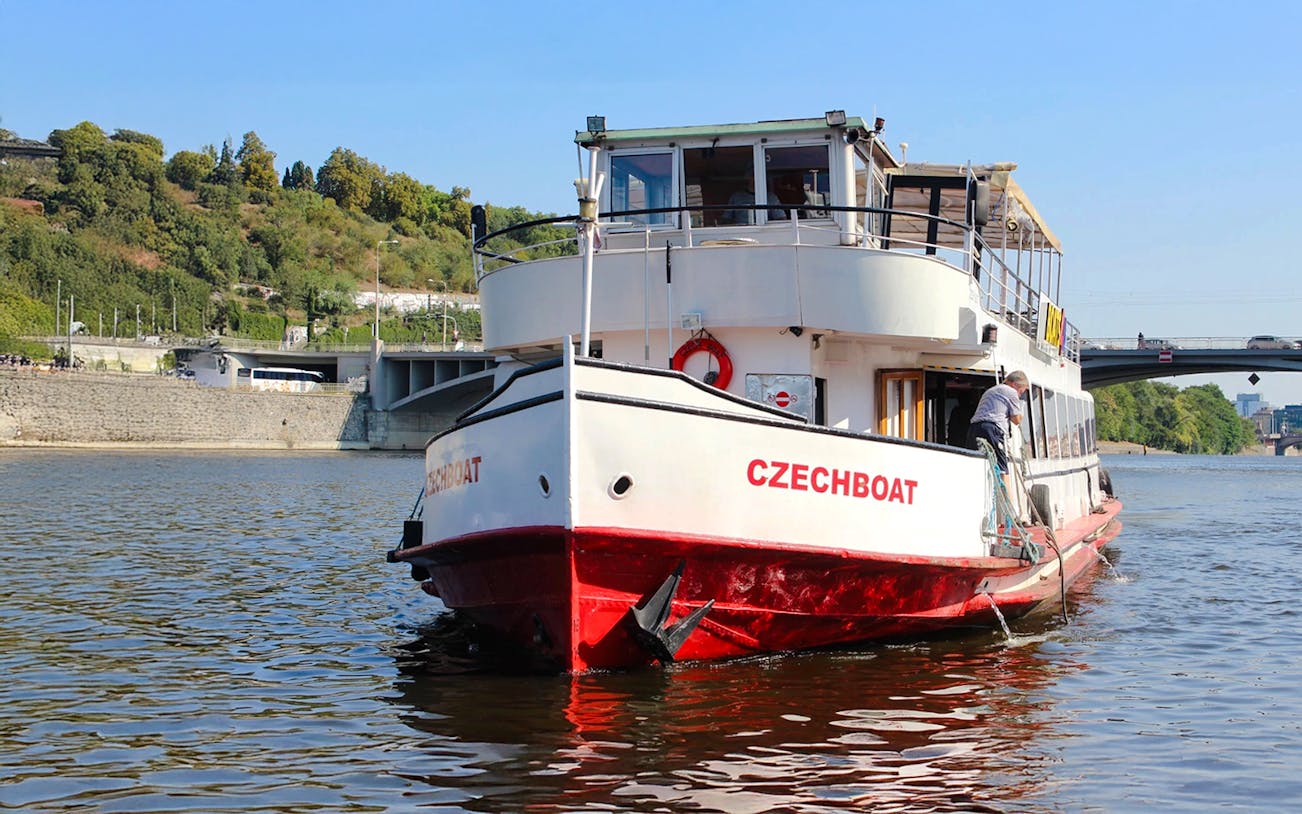 Historic ship Maria Croon on Vltava River, Prague, near a bridge and green hillside.