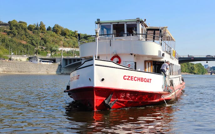 Historic ship Maria Croon on Vltava River, Prague, near a bridge and green hillside.