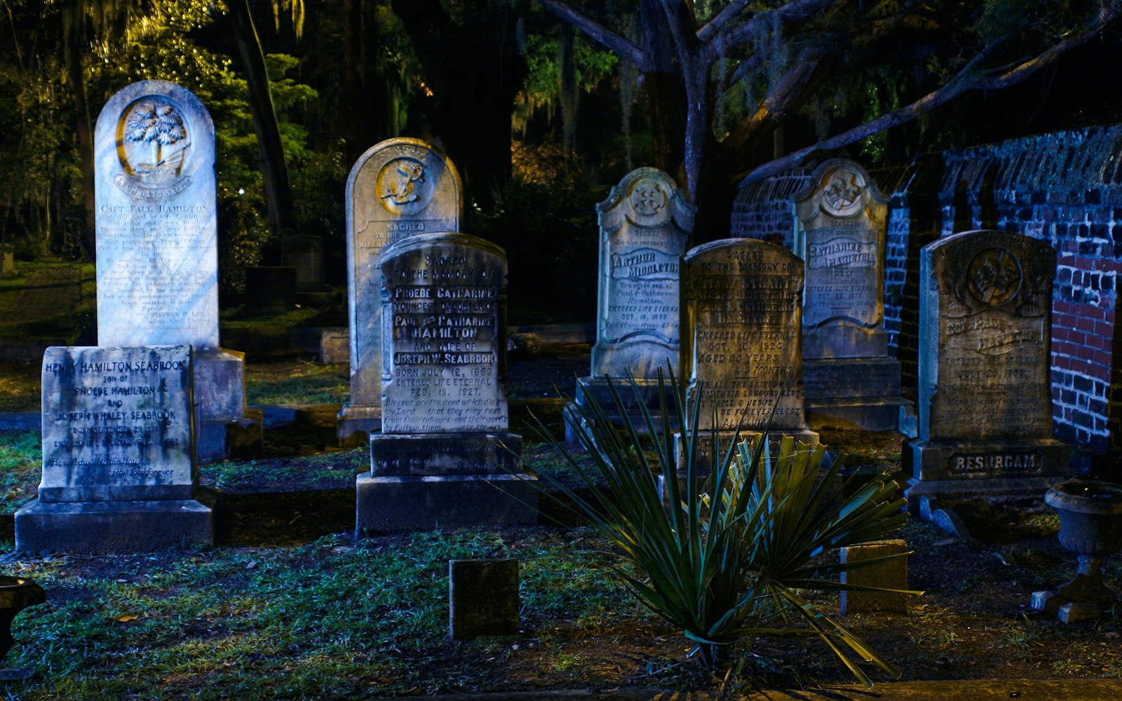 Old gravestones in a dimly lit cemetery at night, surrounded by trees and a brick wall.