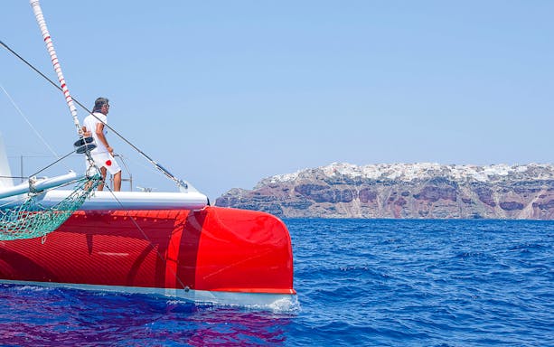 Catamaran sailing near Santorini cliffs during cruise with BBQ and open bar.