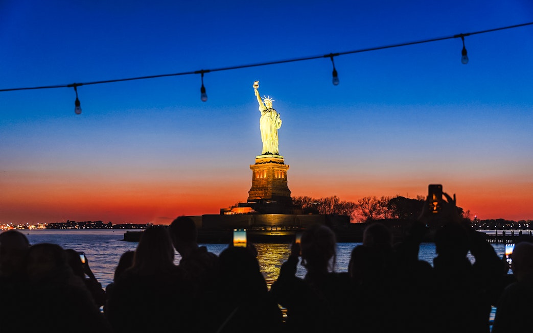 Statue of Liberty illuminated at night with sunset sky, viewed from a boat.