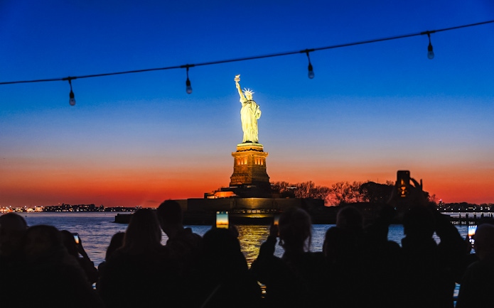 Statue of Liberty illuminated at night with sunset sky, viewed from a boat.