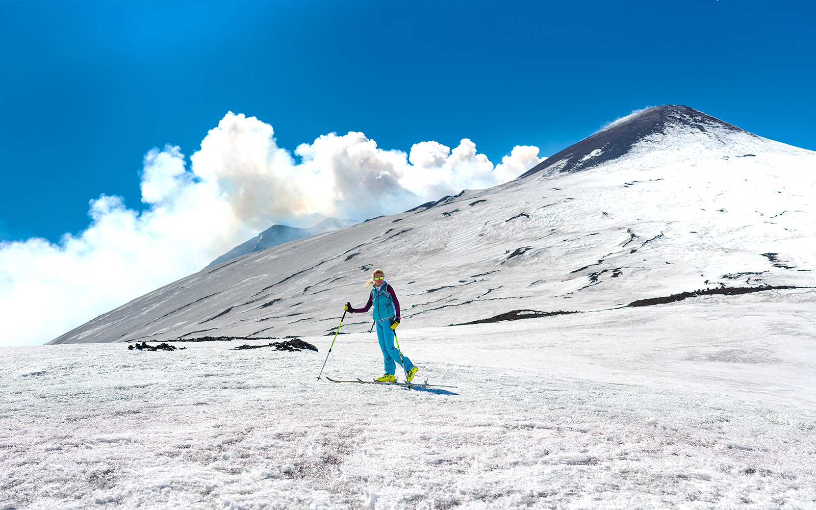 Skiers descending snowy slopes of Mount Etna, Sicily, with volcanic landscape in the background.