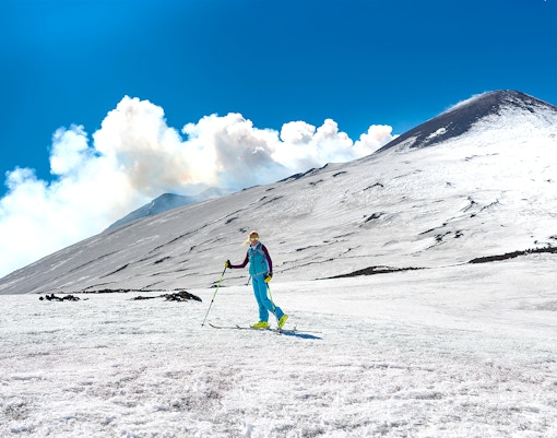 Skiers descending snowy slopes of Mount Etna, Sicily, with volcanic landscape in the background.