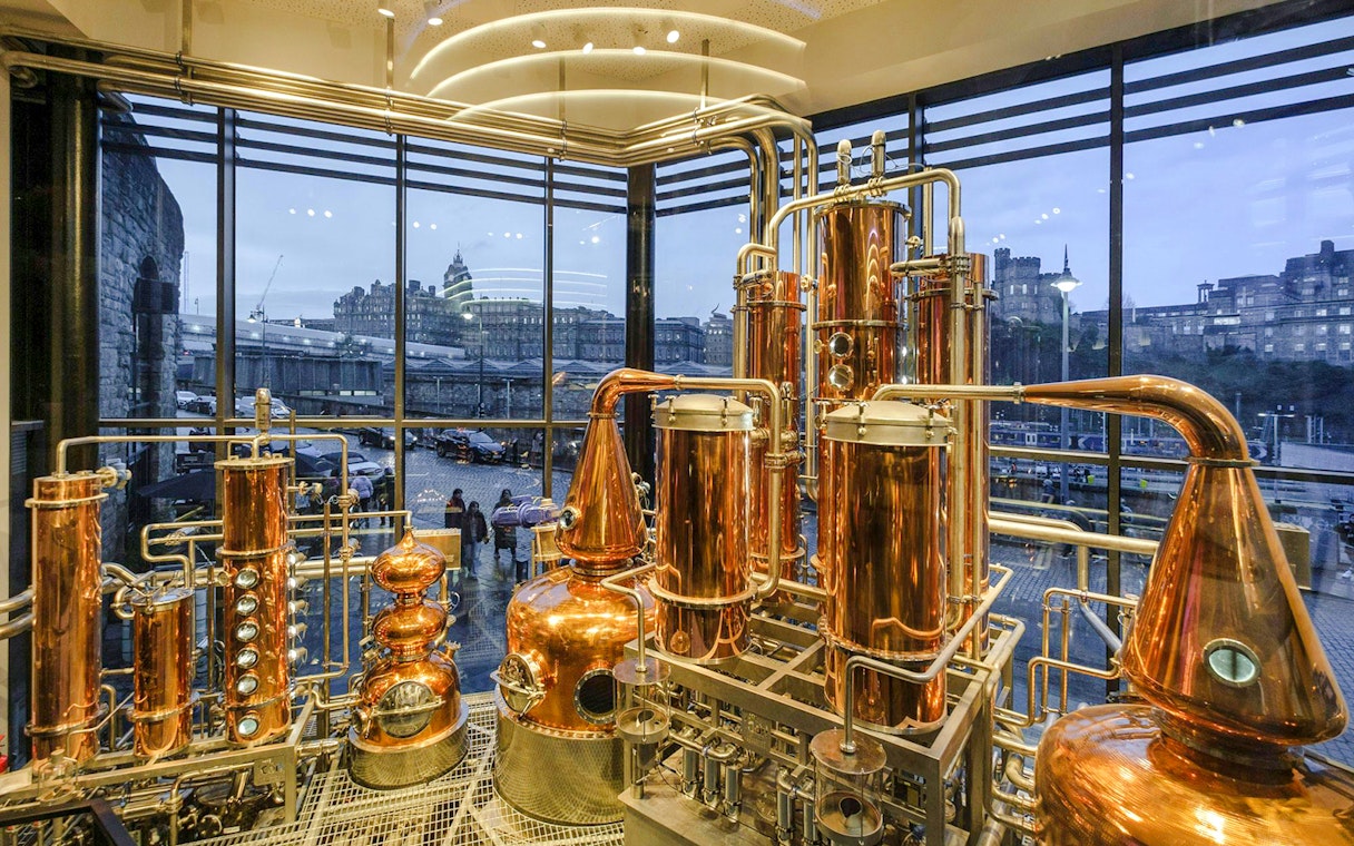 Copper stills inside Edinburgh gin distillery with city view through large windows.