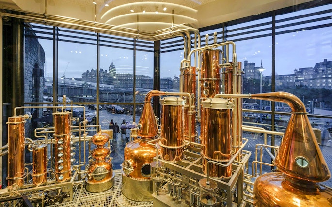 Copper stills inside Edinburgh gin distillery with city view through large windows.