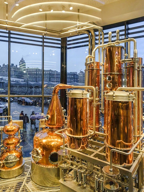 Copper stills inside Edinburgh gin distillery with city view through large windows.