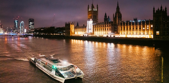 Uber Boat on Thames River at night passing illuminated Houses of Parliament, London.