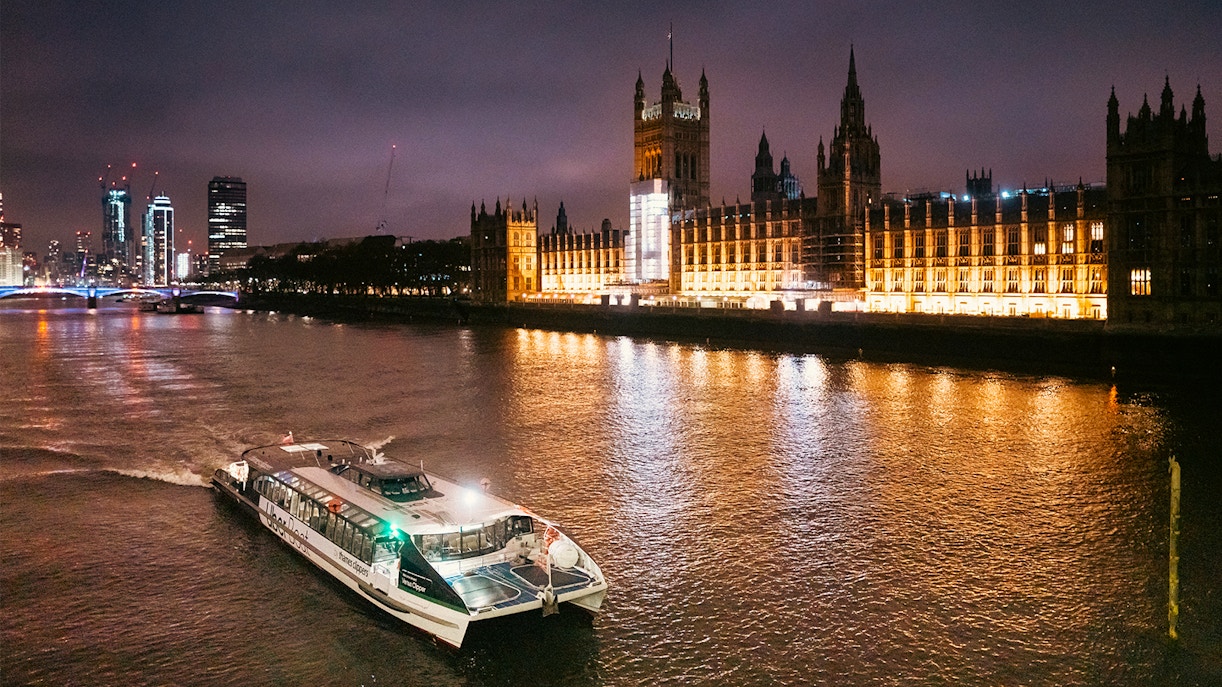 Uber Boat on Thames River at night passing illuminated Houses of Parliament, London.