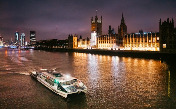 Uber Boat on Thames River at night passing illuminated Houses of Parliament, London.