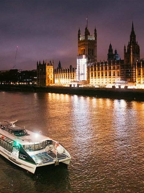 Uber Boat on Thames River at night passing illuminated Houses of Parliament, London.