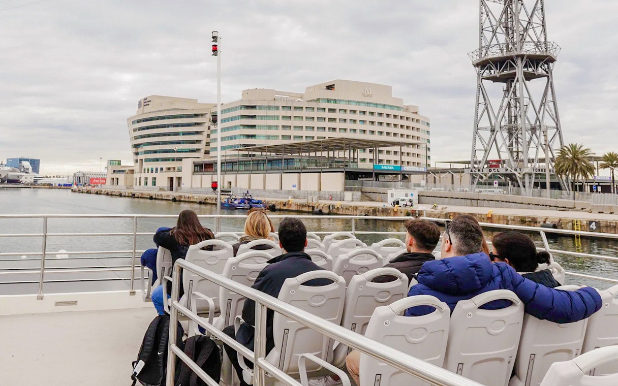 Tourists on a boat tour near a modern building and tower in a harbor.