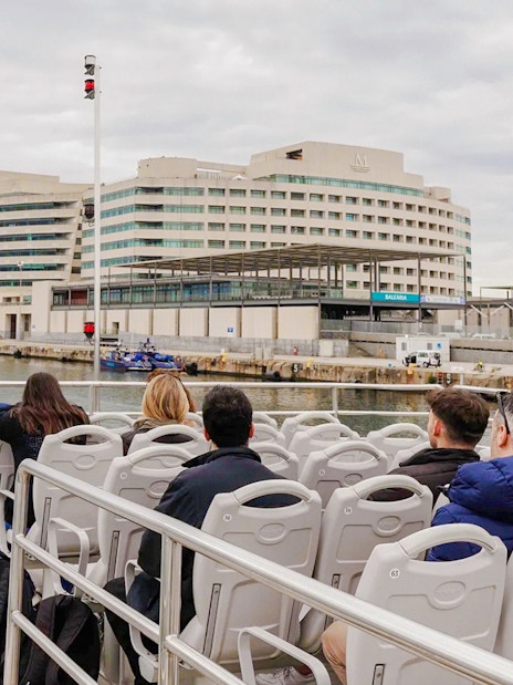 Tourists on a boat tour near a modern building and tower in a harbor.