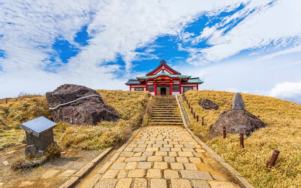 Pathway leading to a red shrine on Mt. Komagatake, surrounded by rocks and grass under a blue sky.