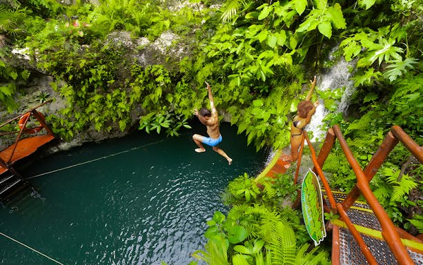 Couple enjoying a jump into a lush cenote surrounded by greenery.
