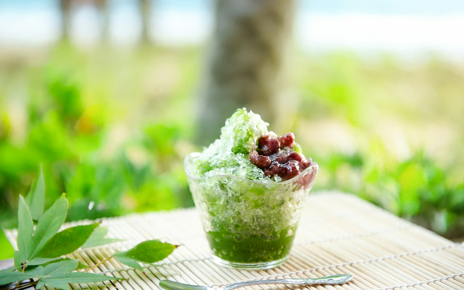 Matcha kakigōri with red beans on a bamboo mat, outdoor setting.