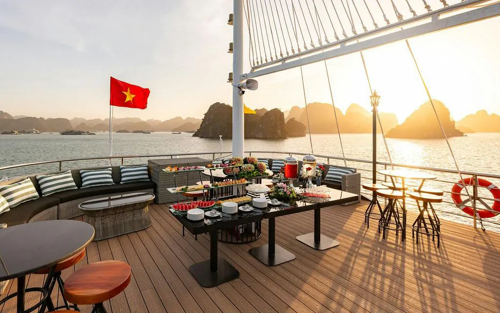 Cruise deck with dining setup overlooking Ha Long Bay at sunset, Vietnam flag visible.
