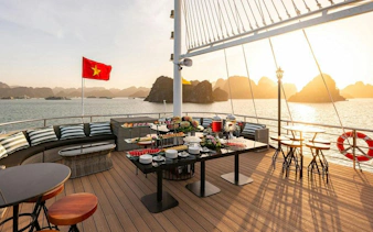 Cruise deck with dining setup overlooking Ha Long Bay at sunset, Vietnam flag visible.