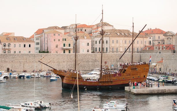 Historic ship docked in Dubrovnik harbor with city walls and buildings in the background.