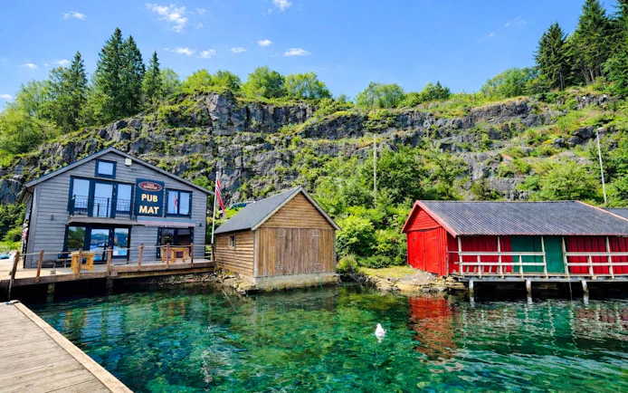 Pub and colorful boathouses by the port in Osterøy, Norway, with rocky hillside backdrop.