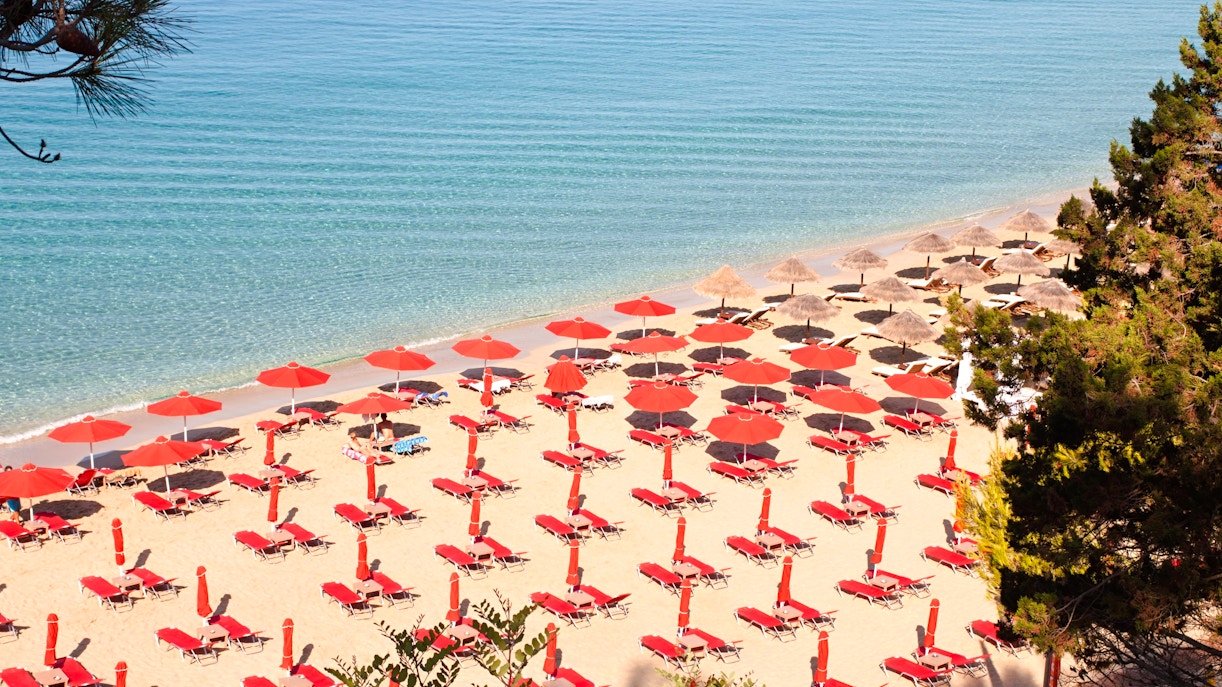Sun loungers and red umbrellas on Makris Gialos beach, Argostoli, Kefalonia, Greece.