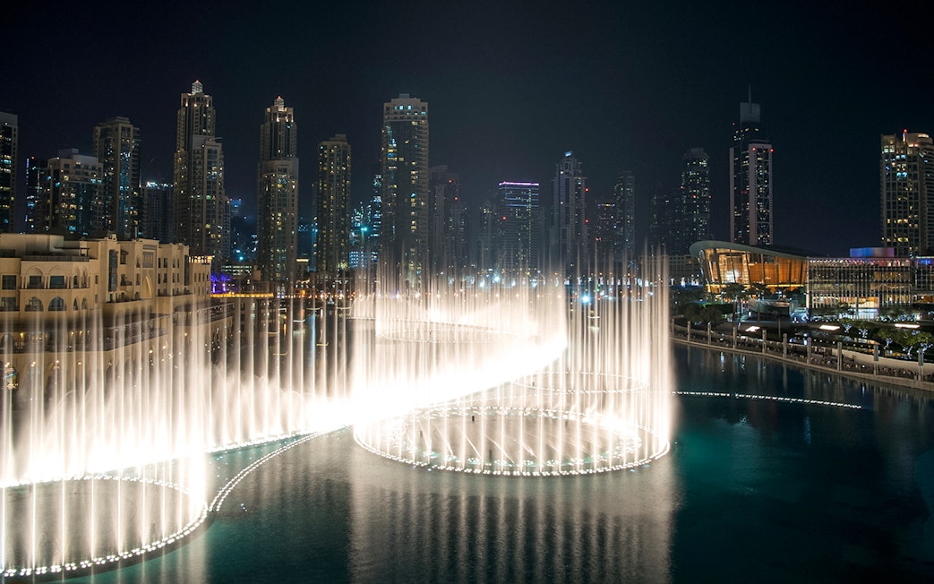 Dubai Fountain Boardwalk with illuminated water jets against city skyline at night.