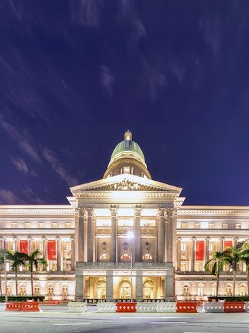 National Gallery Singapore illuminated at night with palm trees in front.