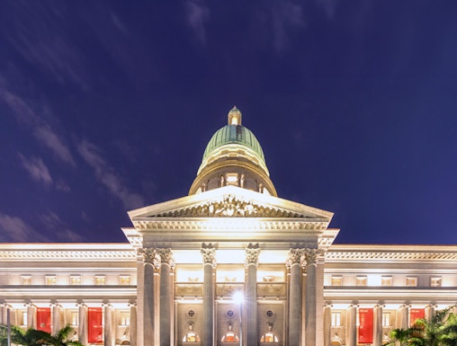 National Gallery Singapore illuminated at night with palm trees in front.