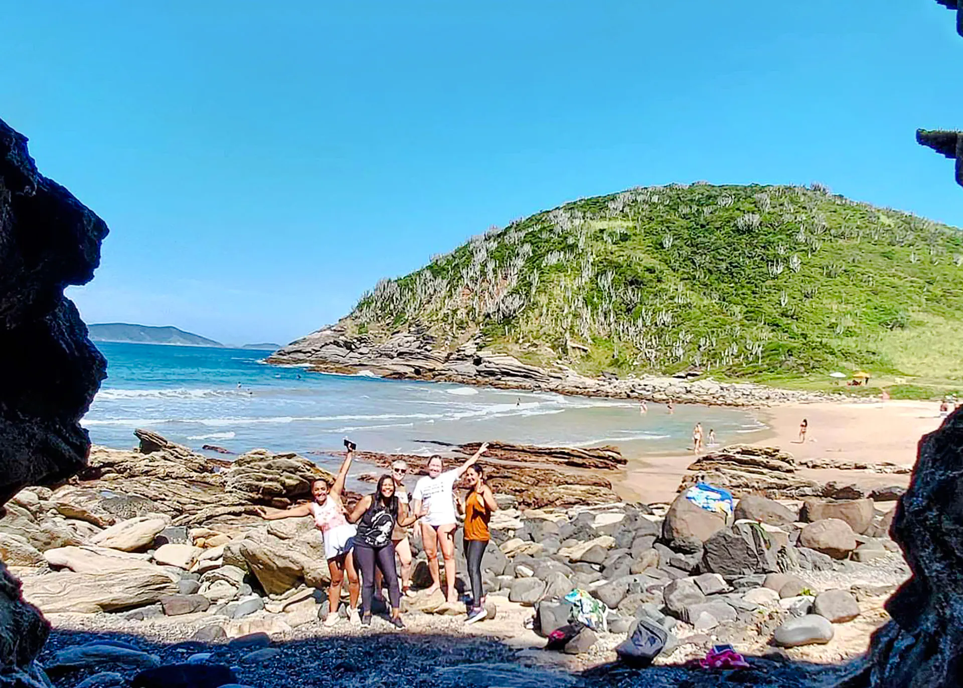 Group exploring rocky beach in Búzios, Rio de Janeiro with lush hills in background.