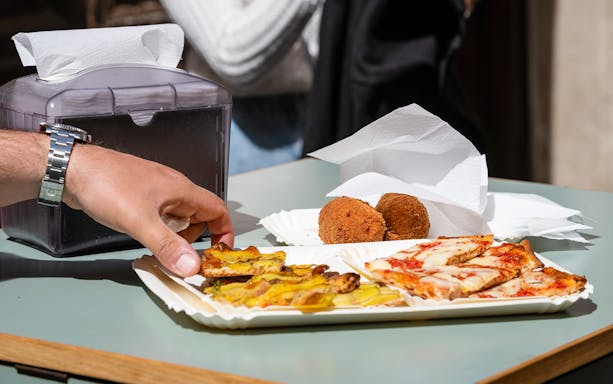 Hand reaching for pizza and fried snacks on a table during a street food tour in Campo de' Fiori, Rome.
